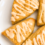 Four golden pumpkin spice scones drizzled with pumpkin latte glaze on a white platter over a marble countertop.