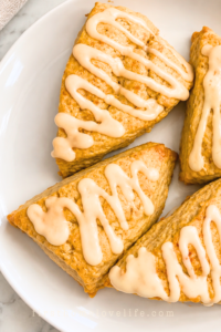 Four golden pumpkin spice scones drizzled with pumpkin latte glaze on a white platter over a marble countertop.
