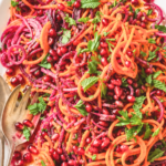 Roasted sweet potato spiral salad with beets, pomegranate seeds, fresh herbs, and pomegranate dressing on a white oval platter.