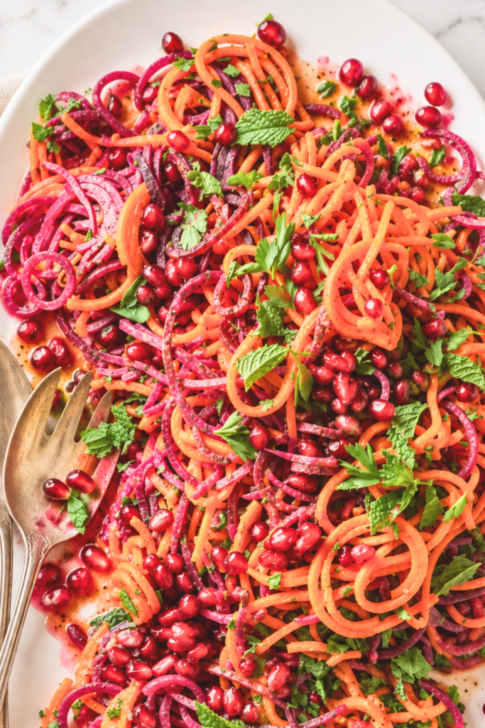 Roasted sweet potato spiral salad with beets, pomegranate seeds, fresh herbs, and pomegranate dressing on a white oval platter.