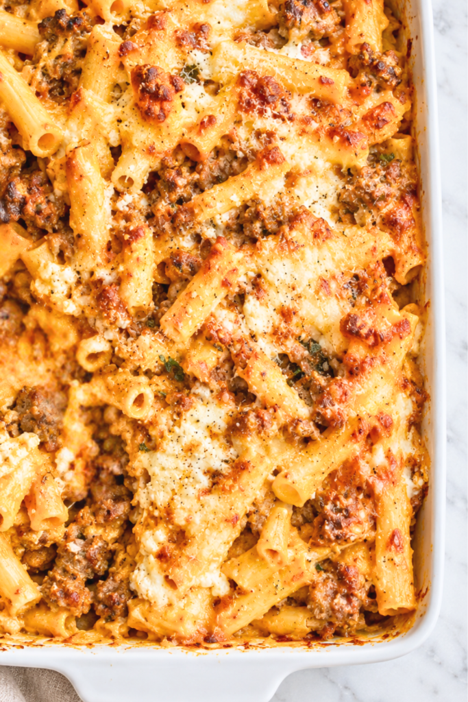 Close-up of sausage and pumpkin baked ziti with browned mozzarella, creamy ricotta, and crumbled Italian sausage in a white baking dish.