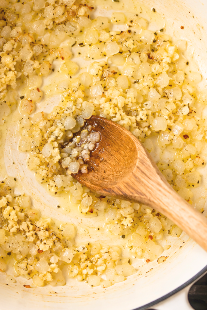 Diced onions and minced garlic sautéing in a white enamel pot with a wooden spoon, viewed from above.
