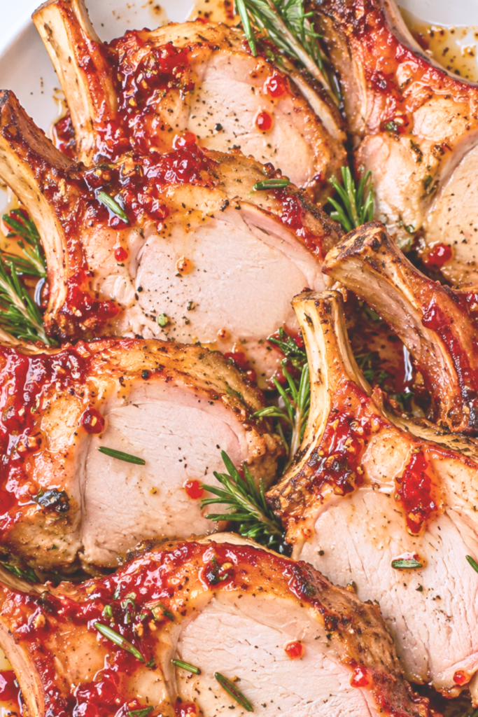 Overhead view of sliced pork crown roast chops brushed with sweet and spicy red pepper jelly glaze and arranged on a white serving platter with rosemary.