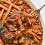 Slow cooker chuck roast with long carrots and thick pomegranate apple cider gravy on a white platter.