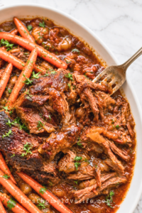 Slow cooker chuck roast with long carrots and thick pomegranate apple cider gravy on a white platter.