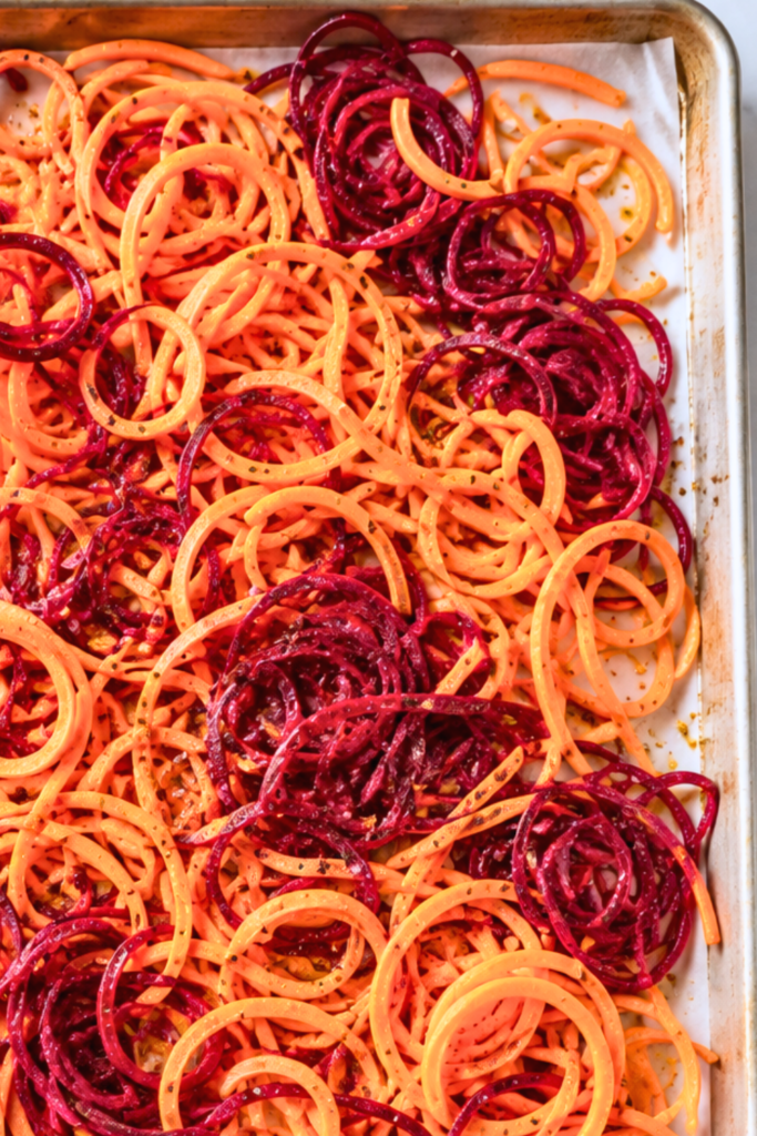 Spiralized sweet potatoes and red beets tossed with olive oil and seasonings on a parchment-lined baking sheet before roasting.